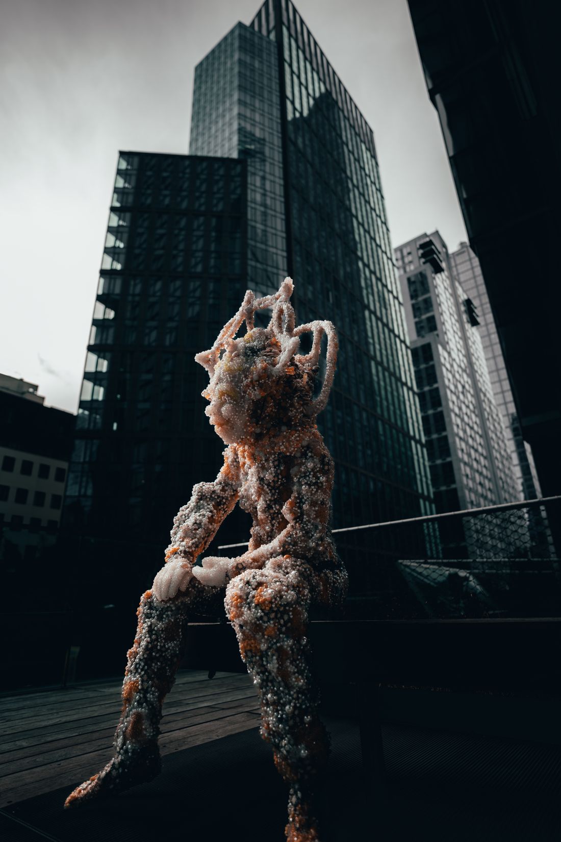 An abstract seated sculpture with a heavily textured surface installed outdoors, glass office towers rising behind it.