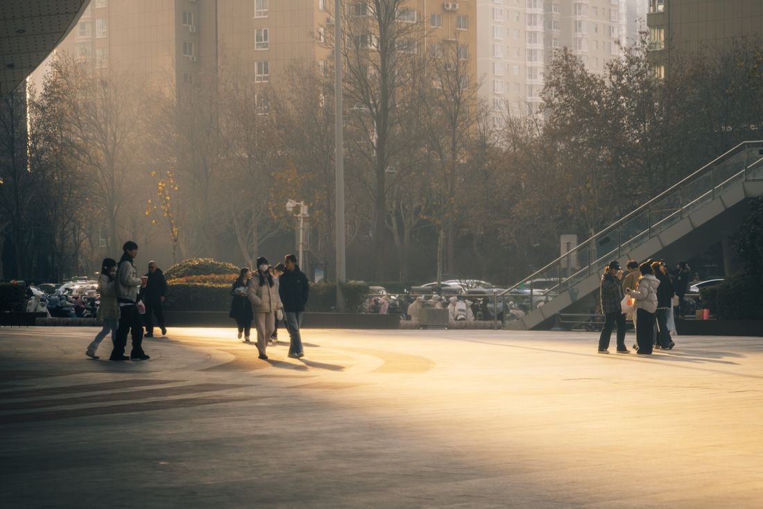 An open city square in low afternoon light with small groups of people walking across the paving and apartment buildings behind.