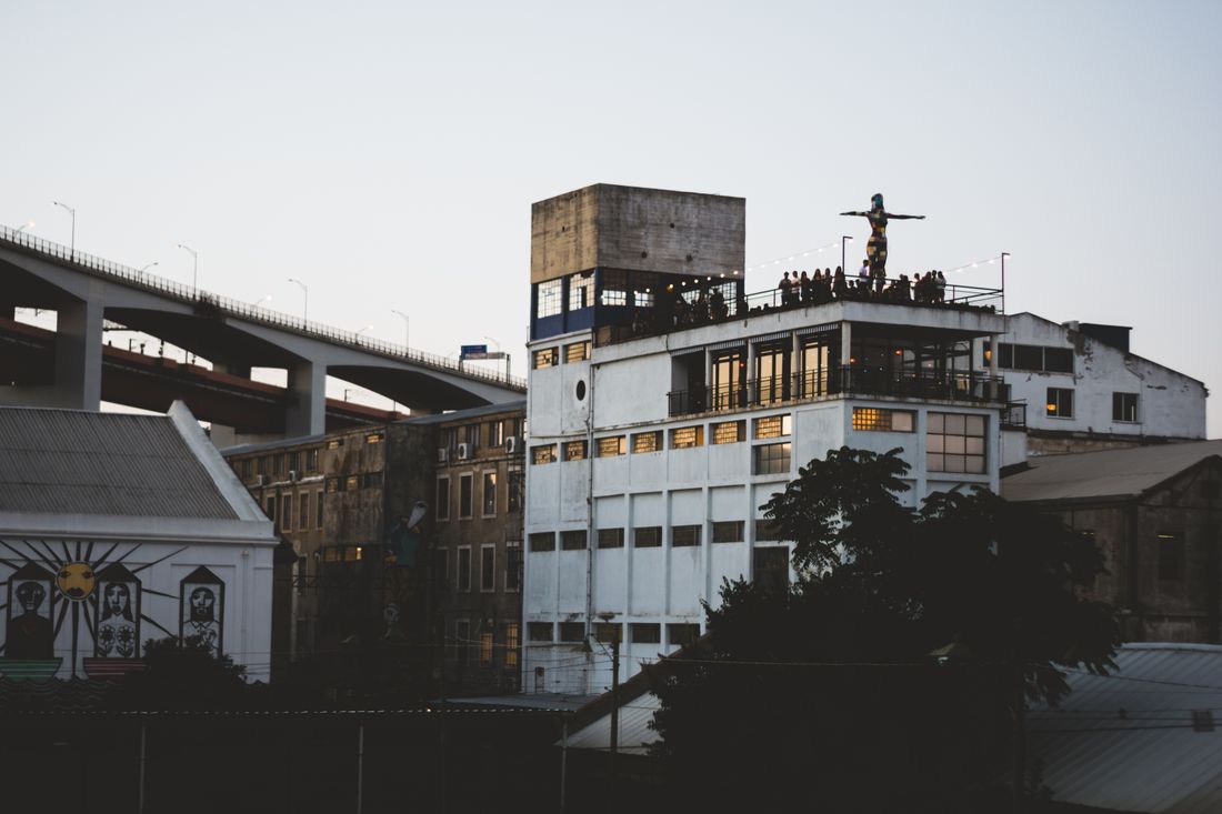 A distant view of a rooftop gathering at dusk, a small crowd standing on the terrace of an industrial building as the sky fades toward evening blue.