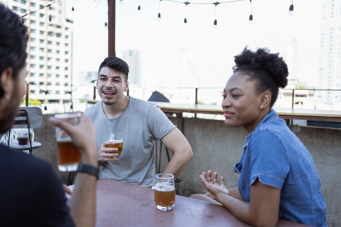 Three people talking and holding drinks at a rooftop table during golden hour, string lights overhead and a city skyline visible behind them.
