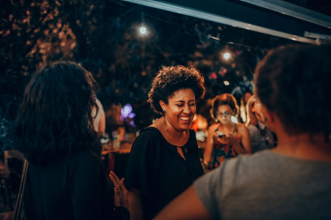 A small group of people talking and laughing outdoors after dark, lit by warm string lights strung overhead.