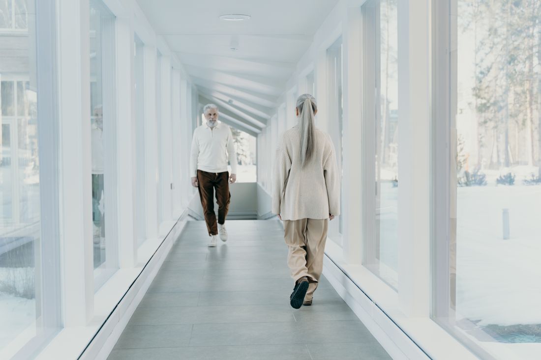 Two people in casual sweaters walking away down a bright, plain glass-walled corridor with a snowy view outside.