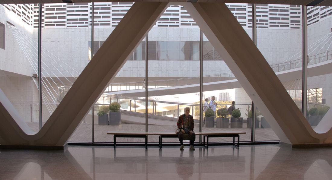 A grand, sunlit museum atrium with dramatic angled beams, a lone visitor seated on a bench and a few others crossing a ramp in the background