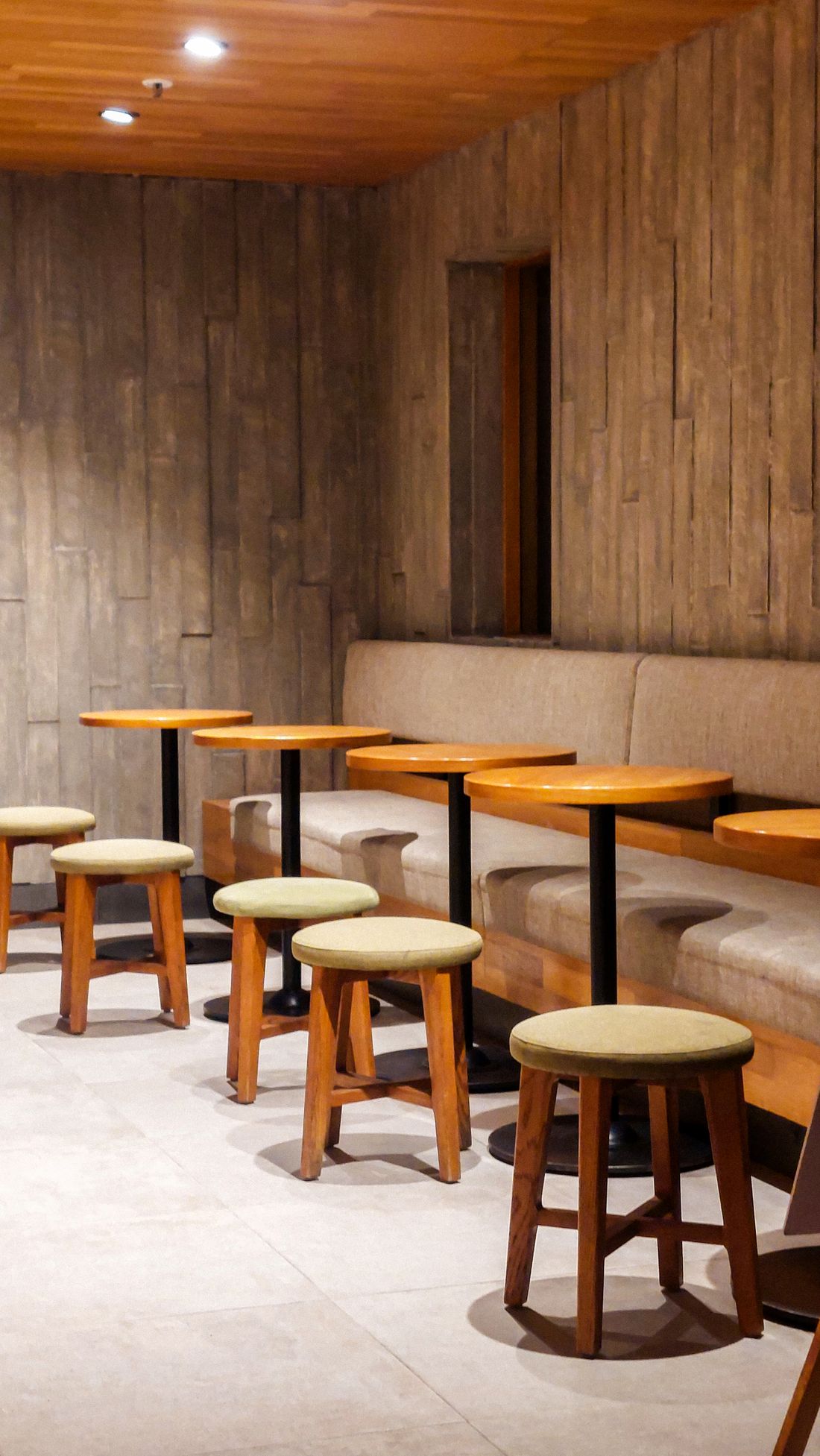An empty lounge seating area with low wooden tables and stools against a wood-paneled wall, no one present