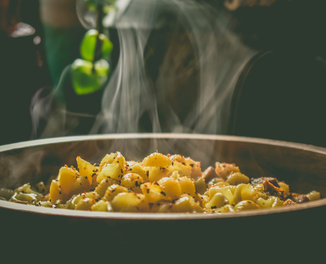 A shallow pan of freshly cooked seasoned pasta shells sends up visible steam against a dark, softly lit background with a hint of greenery.