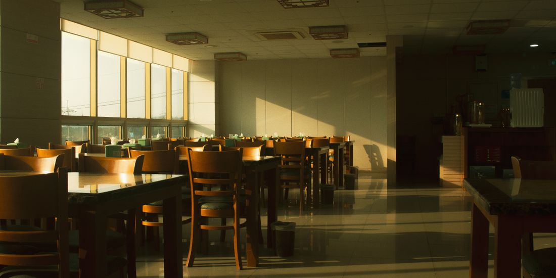 A calm, empty dining room with rows of wooden tables and chairs, warm late-day sunlight streaming through a wall of windows.