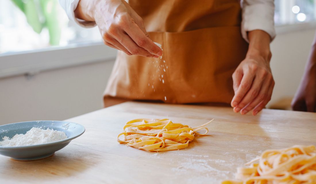 Hands in an apron sprinkle flour over a fresh pile of cut pasta on a wooden prep counter, mid-motion in bright natural window light.
