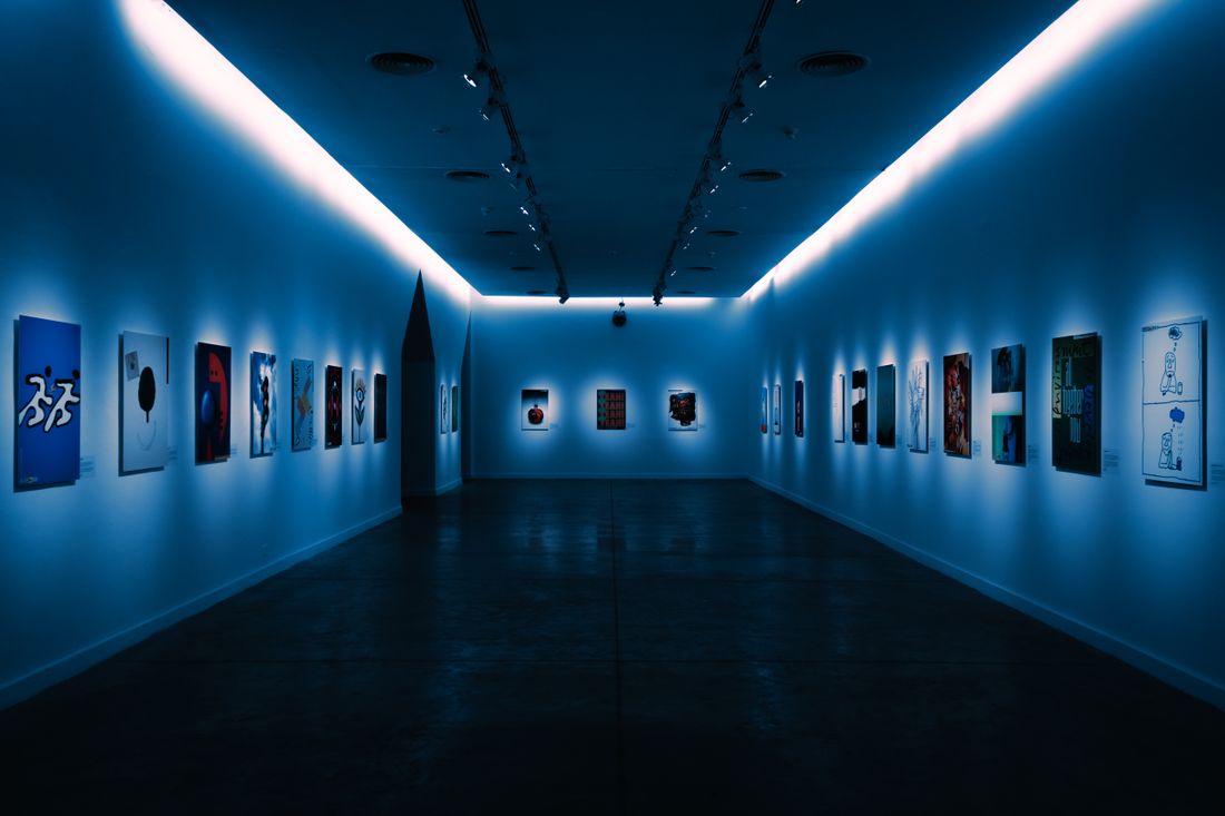 A dimly lit gallery corridor with small framed works on both walls, illuminated by a glowing blue-white cove light along the ceiling, no people in the room.