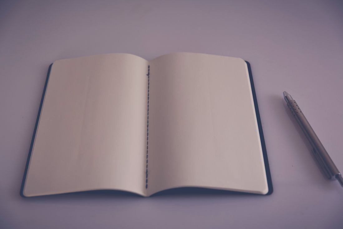 An open notebook with blank pages and a single pen resting beside it on a plain surface, no text, camera, or people visible.