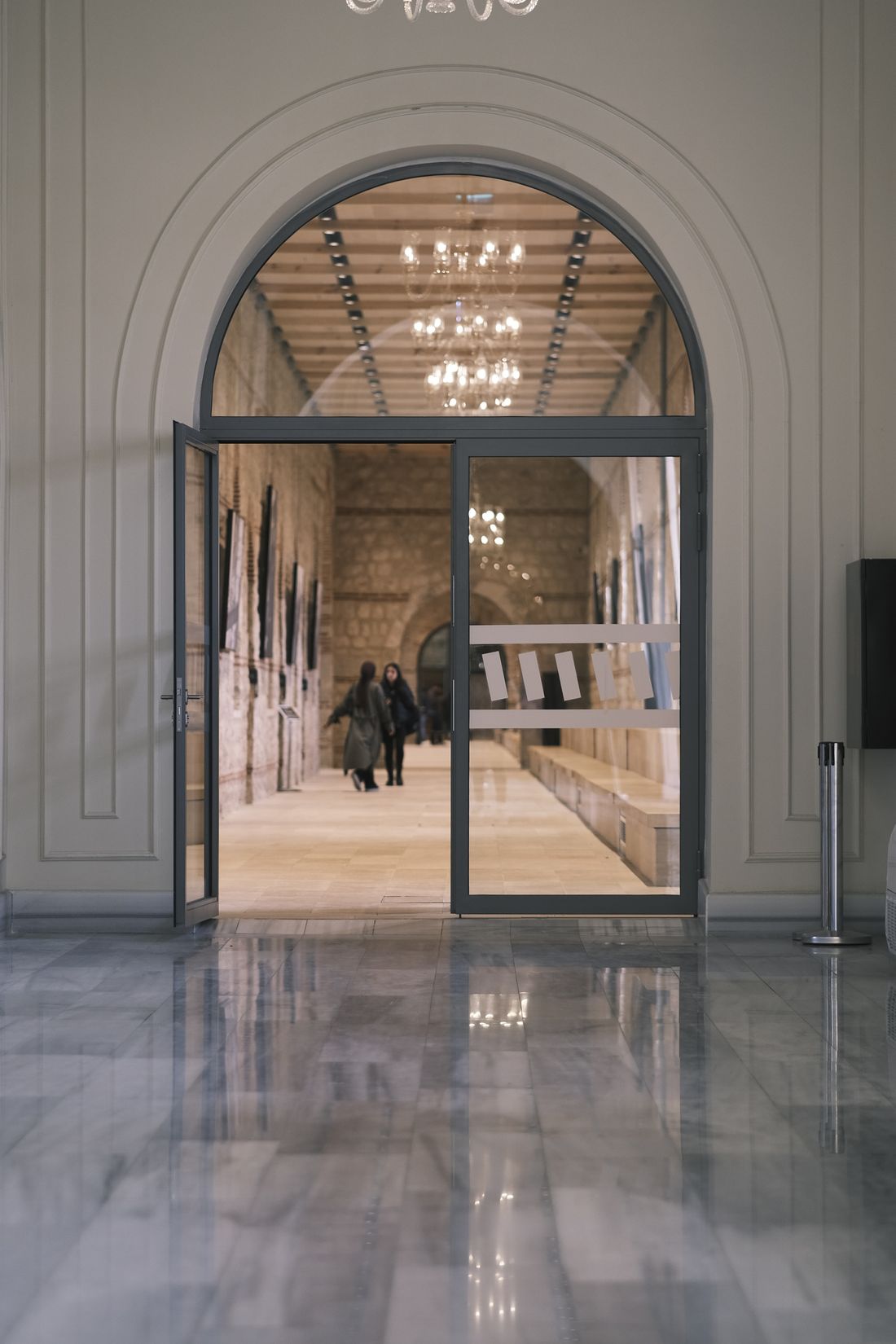 An arched glass doorway framing a warmly lit stone corridor with chandeliers, two people walking inside