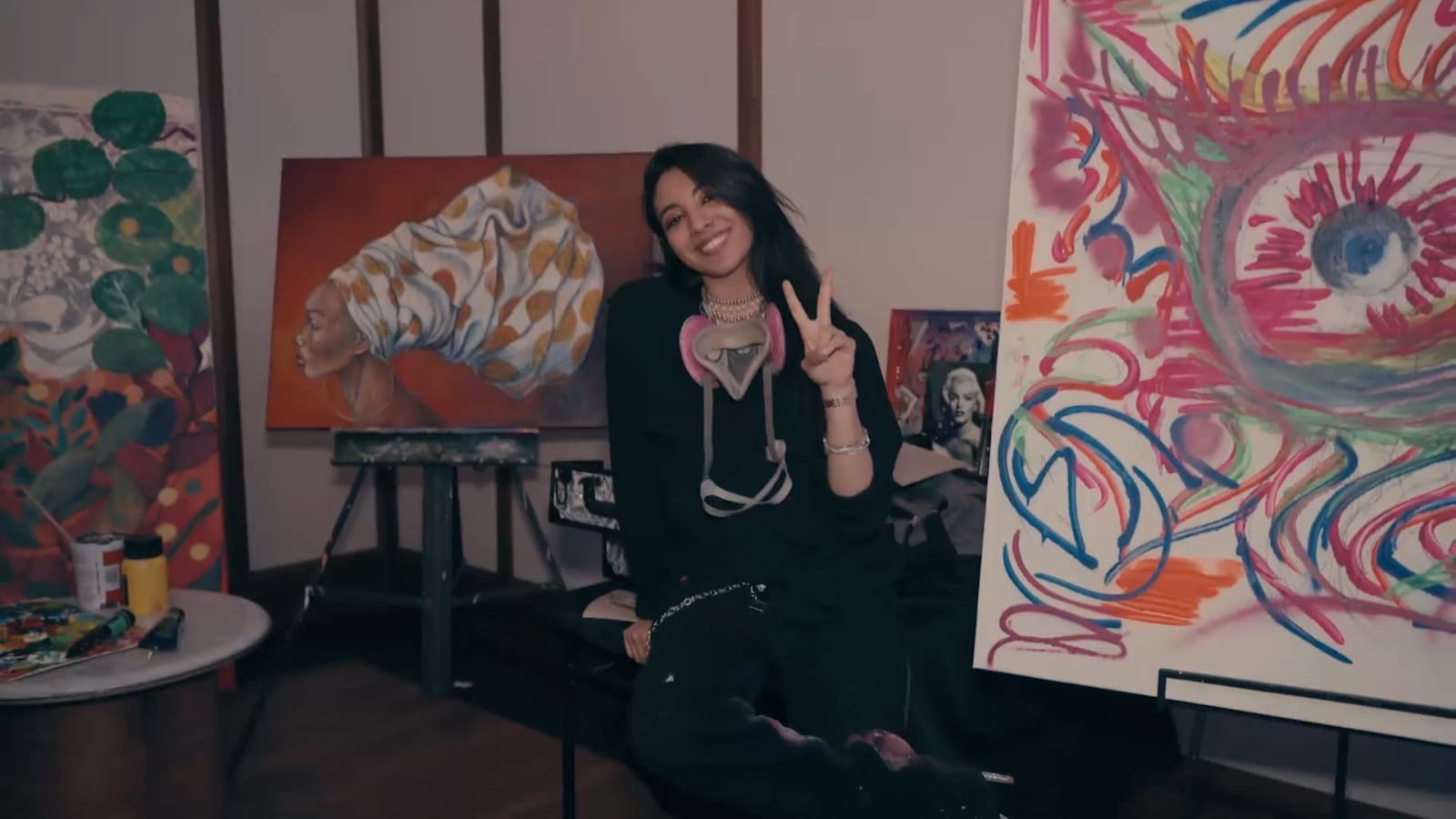A painter sitting among her own canvases in a working studio, colorful abstract and figurative paintings filling the walls around her, an easel in front.