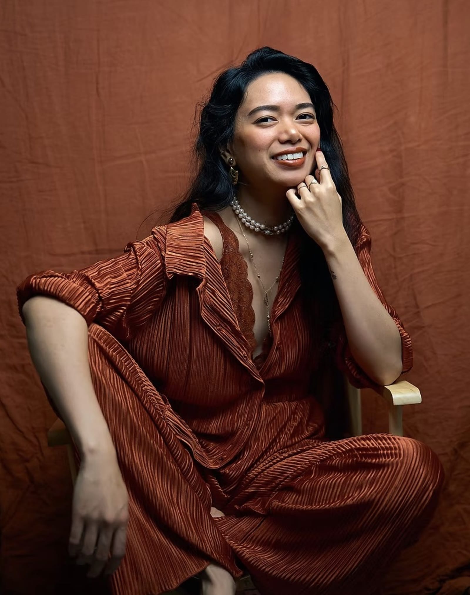 A seated woman in a rust-toned pleated outfit smiling with her hand at her chin, against a plain rust fabric backdrop, no artwork in frame.