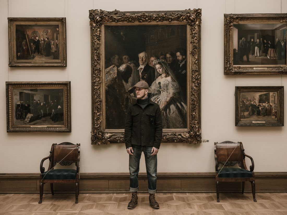 A man in a dark coat standing small in front of a large, ornately framed classical painting on a gallery wall