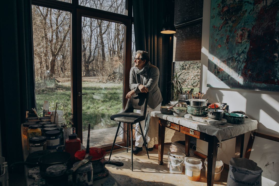 An artist seated near a window in a sunlit studio, with a large painting and work materials visible in the background