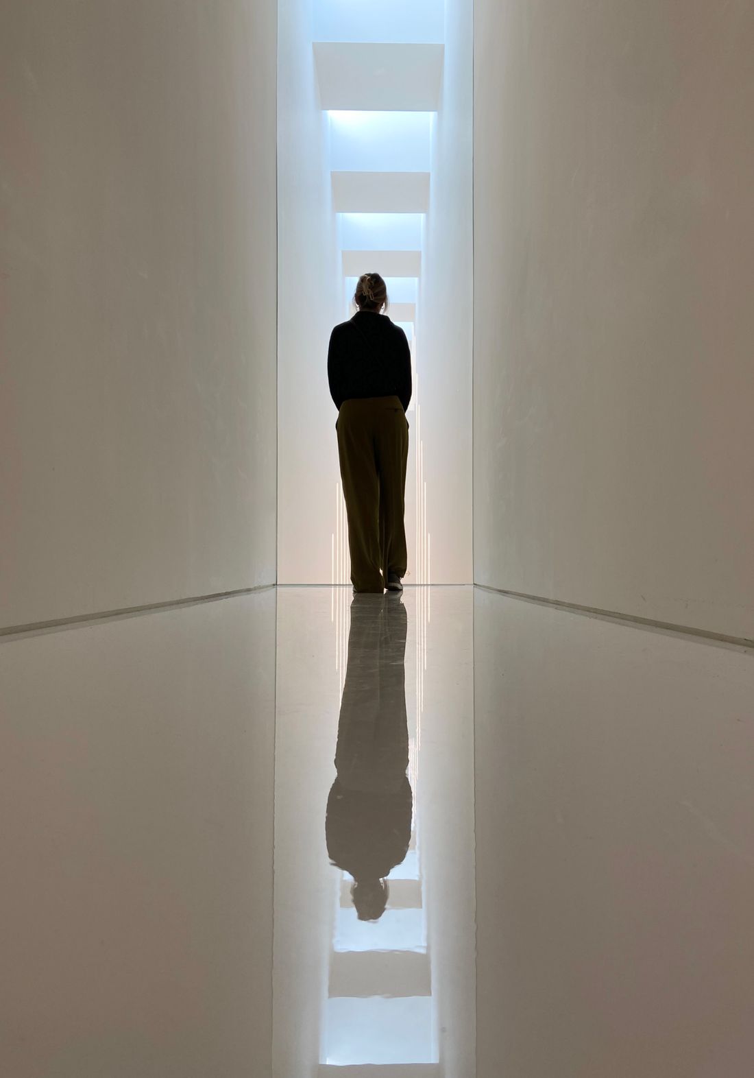 A single figure standing far down a long, narrow, brightly lit corridor, their reflection visible on the polished floor