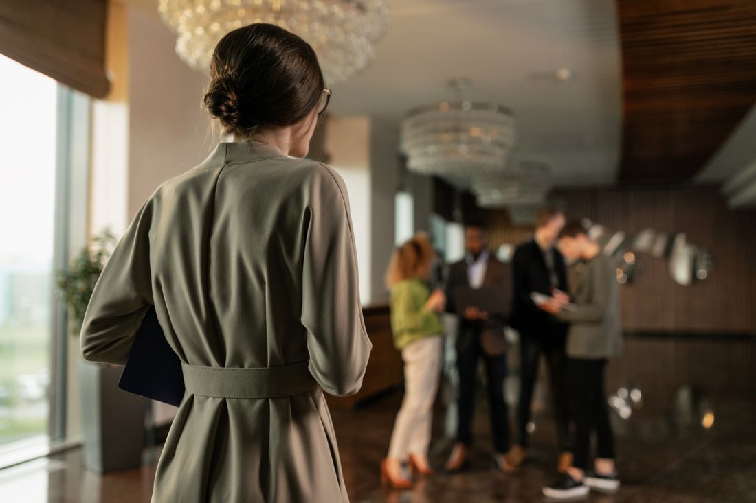 A small group of people in conversation near a lobby reception desk, with open floor space around them and a figure standing in the foreground