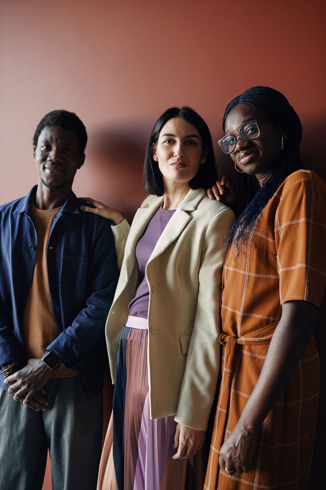 Three people standing close together against a plain colored wall, smiling and facing the camera