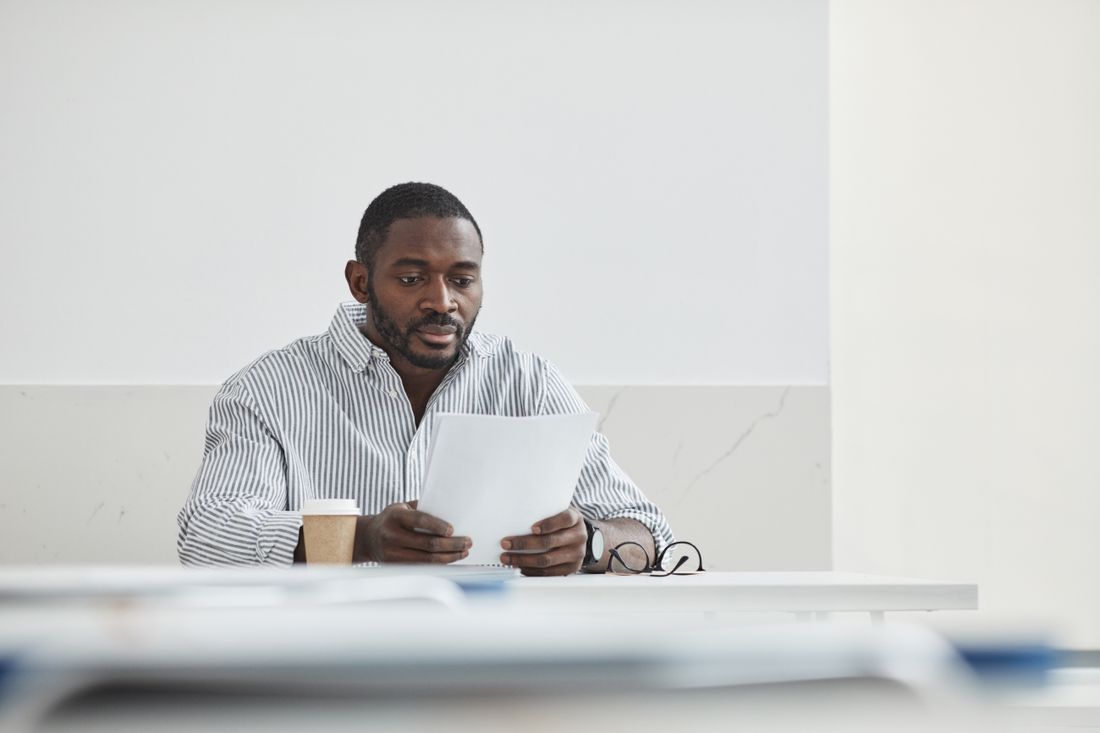 A man seated at a plain desk reading a sheet of paper, a coffee cup and folders nearby.