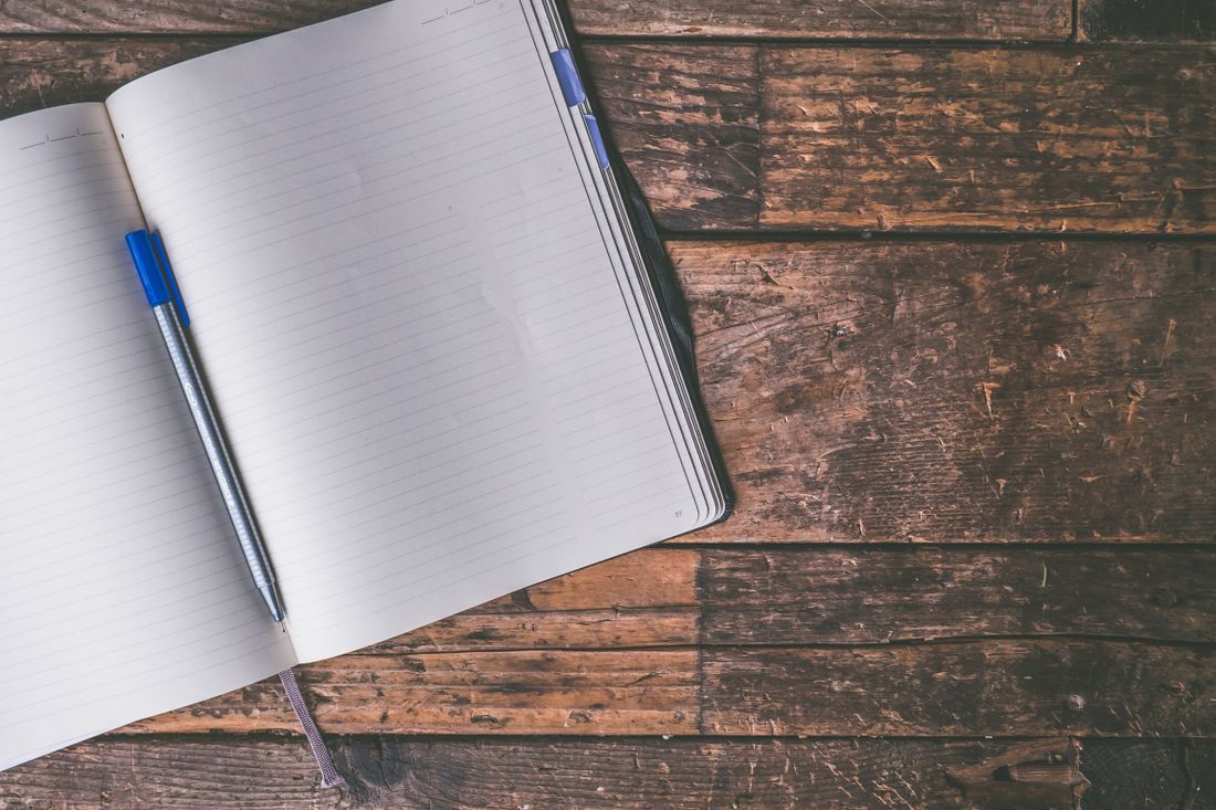 An open blank notebook with a pen resting across the page on a plain wooden table, lit by soft natural light.