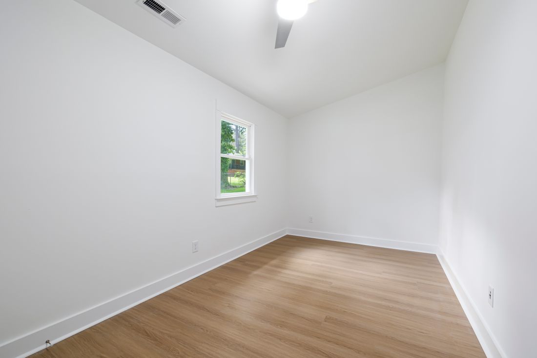 An empty room with plain white walls and wood flooring, lit by daylight from a single window, no furniture or artwork.