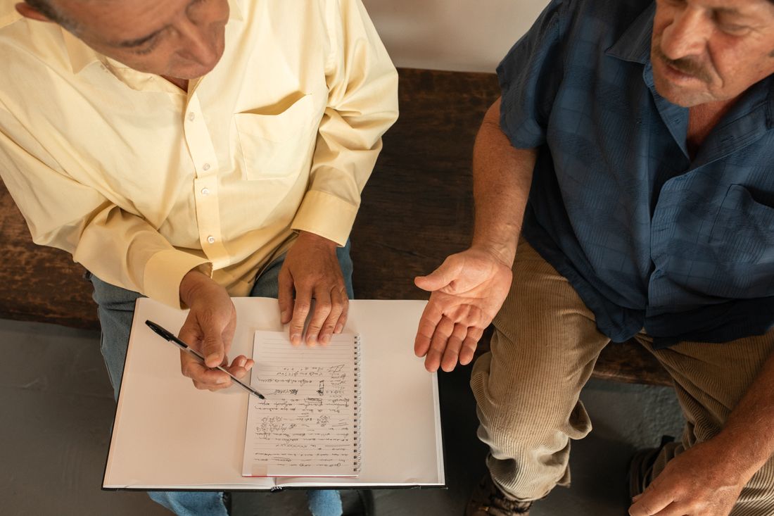An overhead view of two people in plain button-up shirts looking at a handwritten spiral notepad with a pen, seated on a wooden bench.