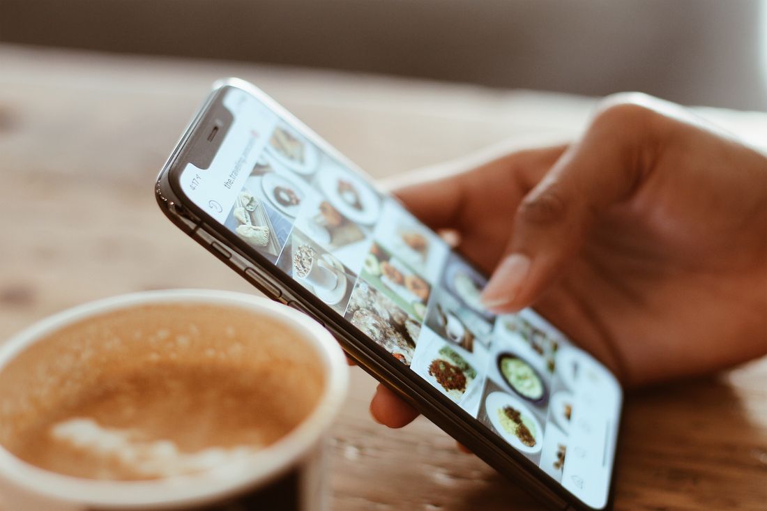 A hand holding a smartphone showing a grid of photos while browsing next to a cup of coffee