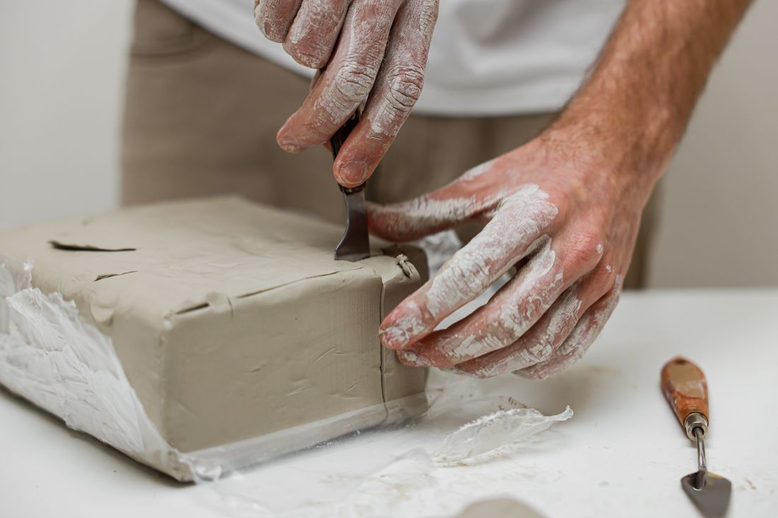 Close-up of hands shaping and cutting a block of clay with a tool on a studio table