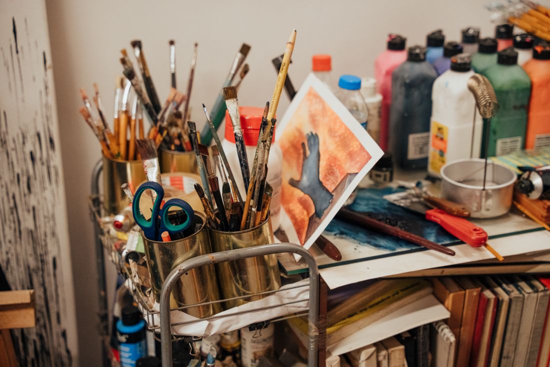 A colorful artist studio corner with jars of brushes, paint tubes, and a work in progress