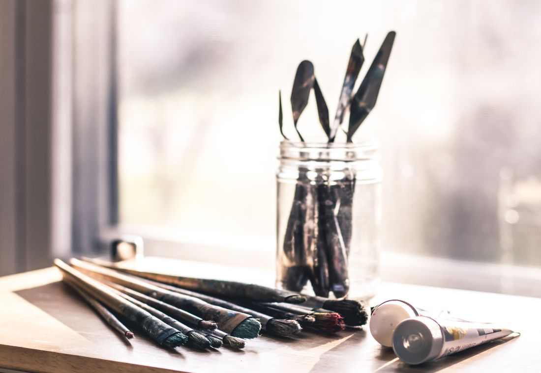 A jar of paintbrushes and a tube of paint on a sunlit windowsill table in a studio