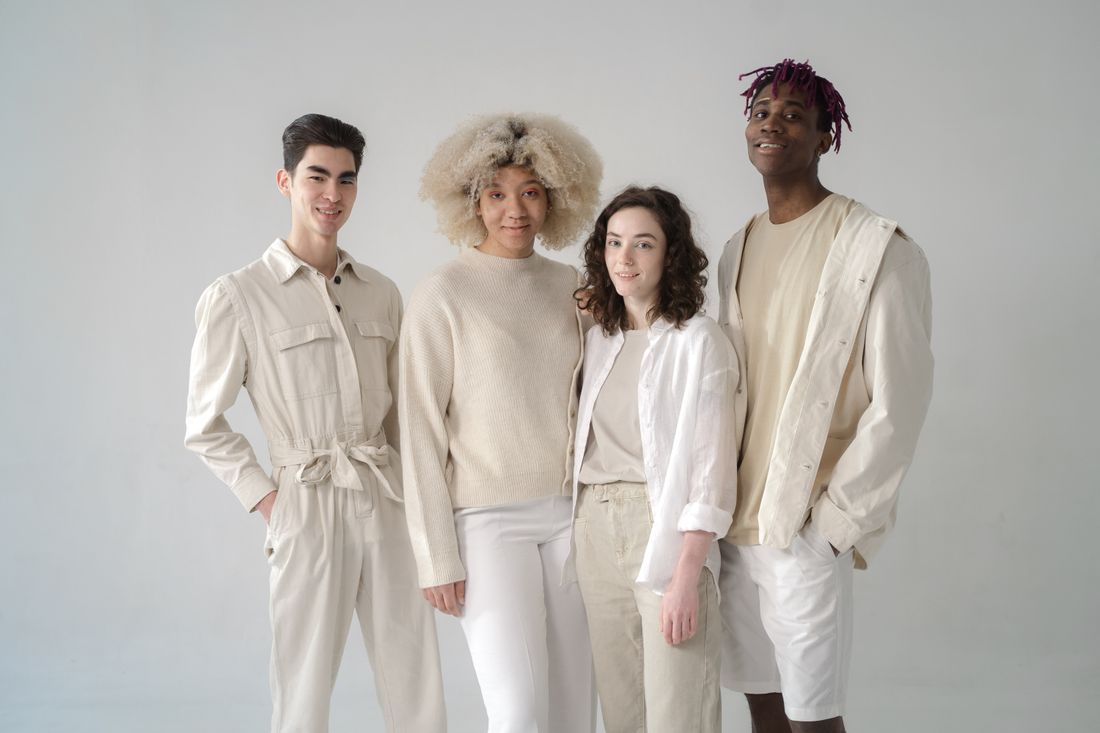 Four adults standing together against a plain wall, each in a different garment but all in similar cream and beige tones.
