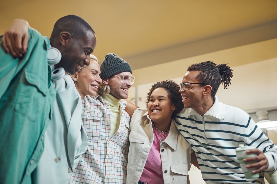 Five adults in different casual outfits standing close together with arms around each other, laughing.