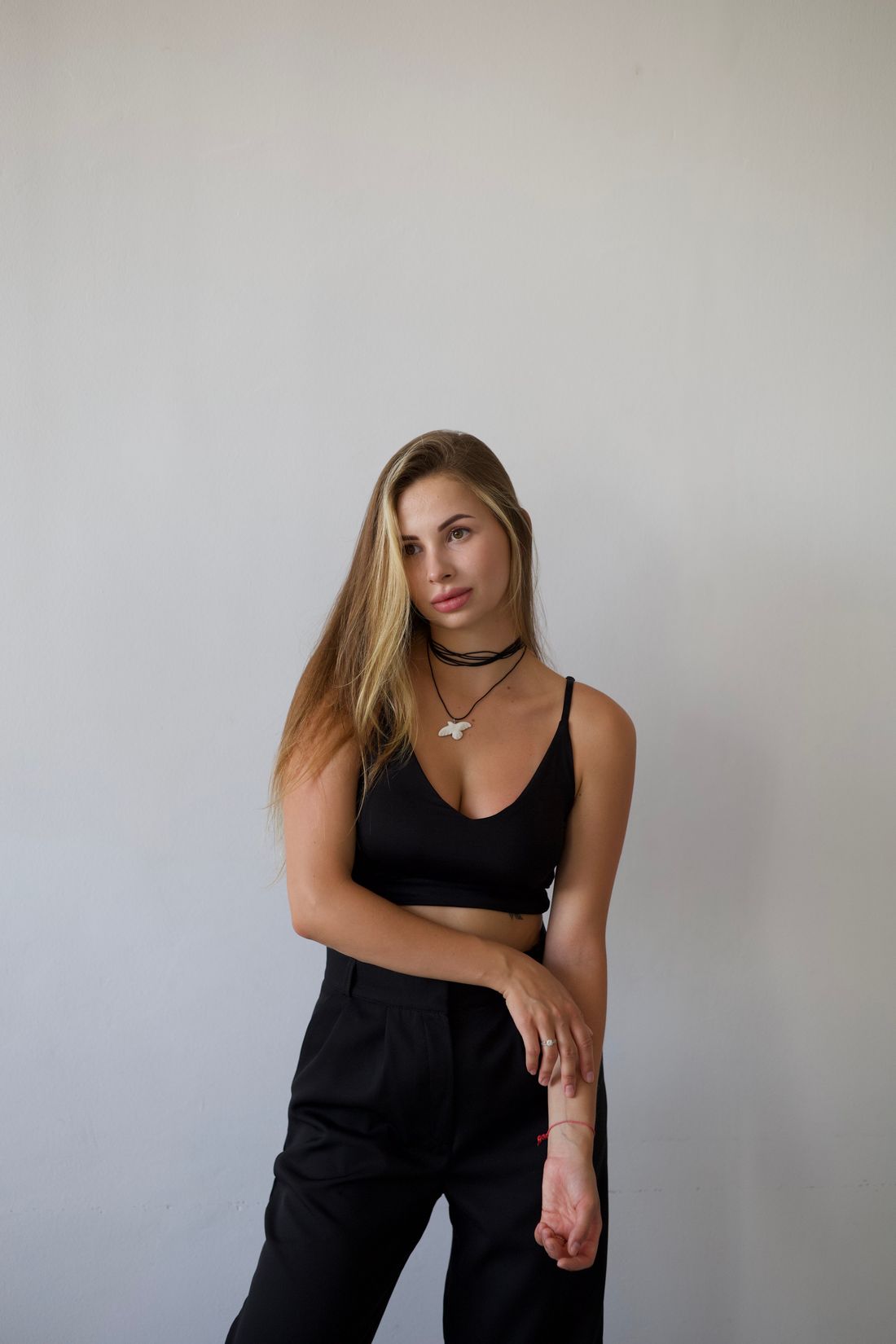 Portrait of a person in a black outfit standing against a plain light wall and wearing layered necklaces.
