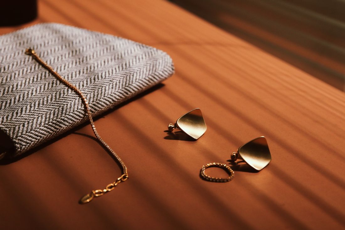 Unbranded gold necklace, earrings, and ring arranged beside a textured clutch on a sunlit wooden surface.