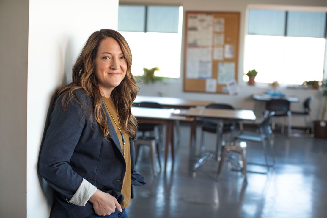 A woman in a navy blazer leans against a plain white wall, smiling, in a bright room with tall windows