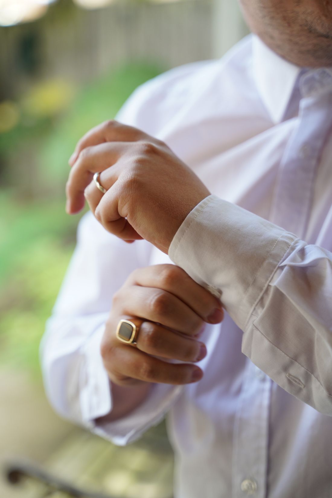 Close detail of hands buttoning a white shirt cuff, with a plain gold ring, against a softly blurred outdoor background