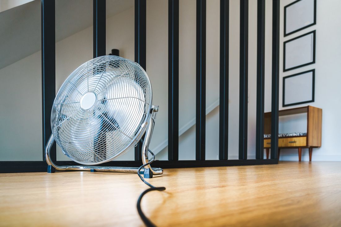 A chrome pedestal fan running on a wood floor in a bright room, positioned near a staircase and a small console table