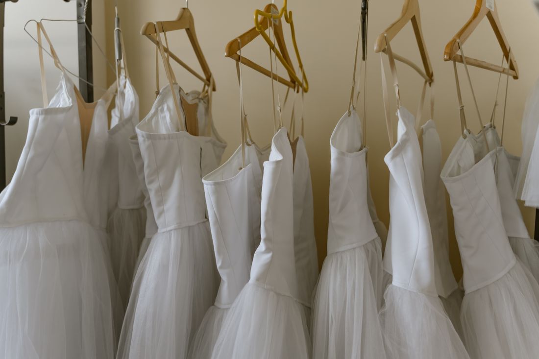 A row of near-identical white dresses hanging on wooden and metal hangers on a wardrobe rack, seen from a slight distance against a plain wall.