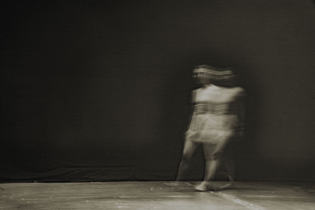 A motion-blurred, unrecognizable figure walking mid-stride across a dark stage floor, clothing visibly in motion, face obscured by blur.