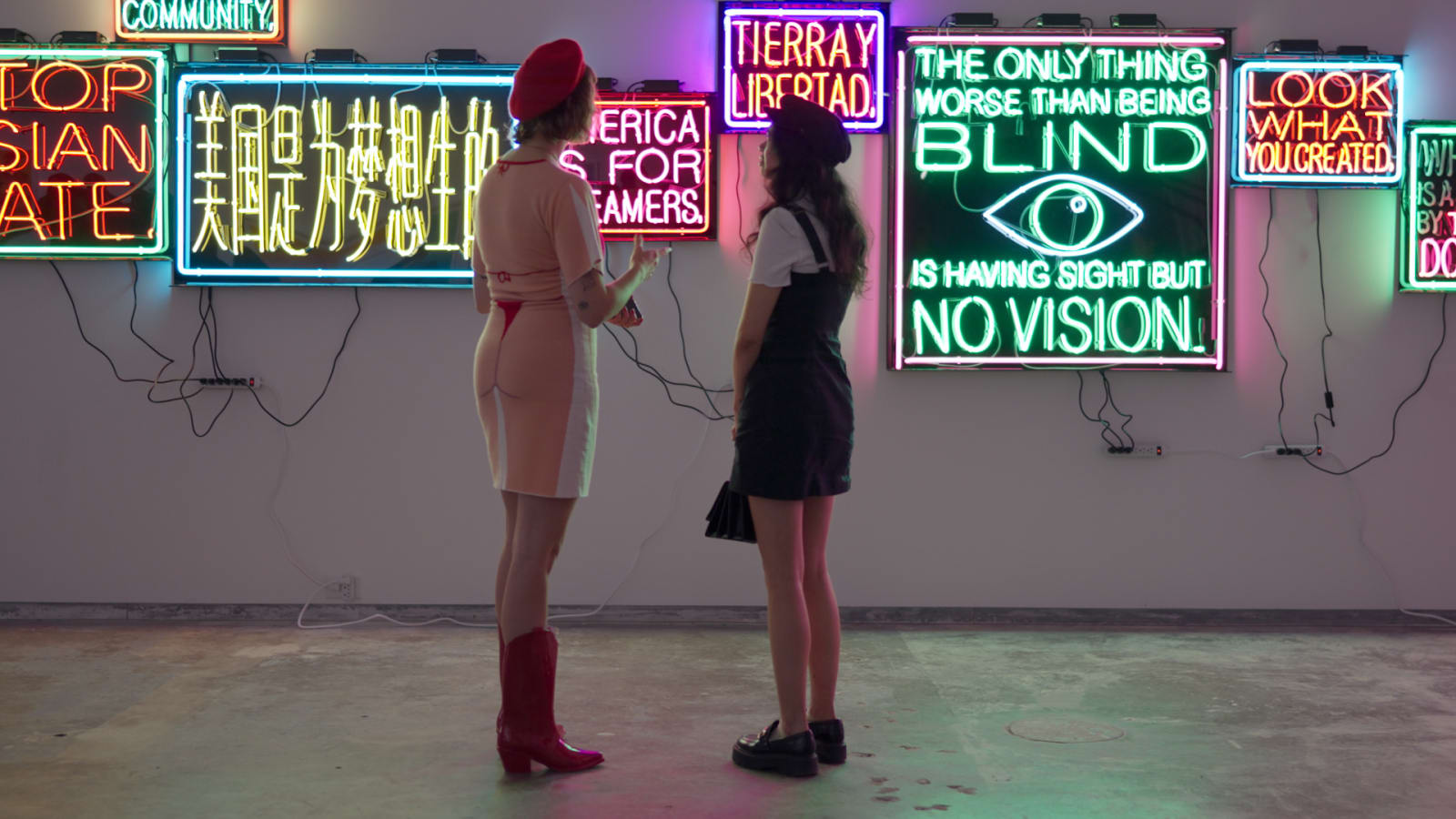 Two gallery visitors in red berets and boots, seen from behind, standing in front of a wall covered in colorful neon text artworks