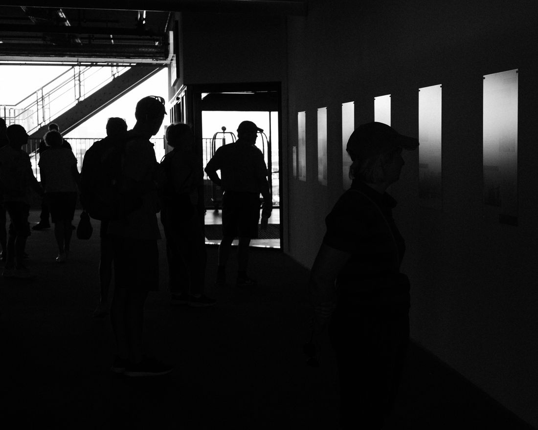 Silhouettes of gallery visitors standing in a darkened hallway with framed works lit along one wall
