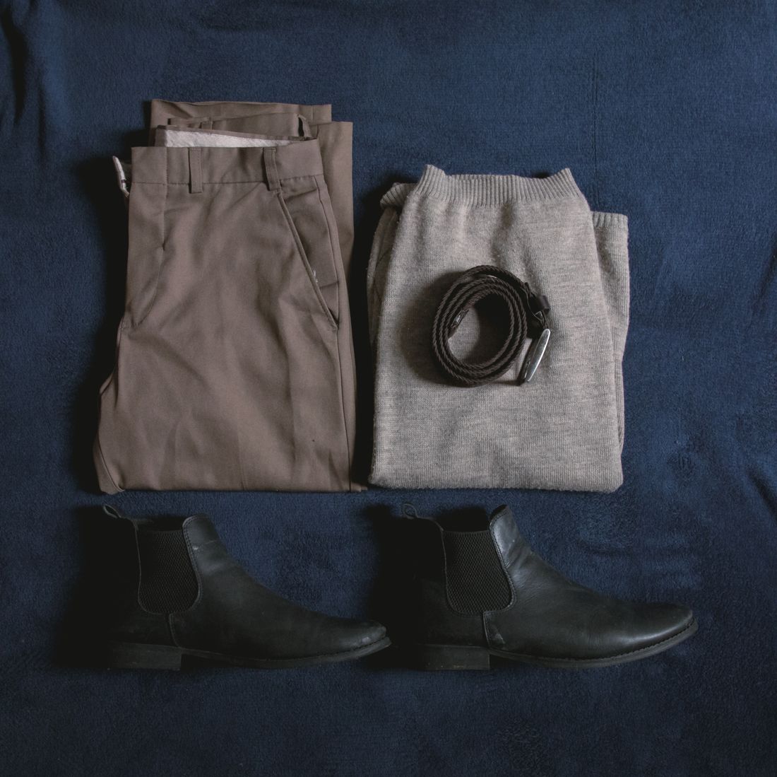 Flat lay of folded trousers, a gray sweater, a leather belt, and black boots on a dark blue background