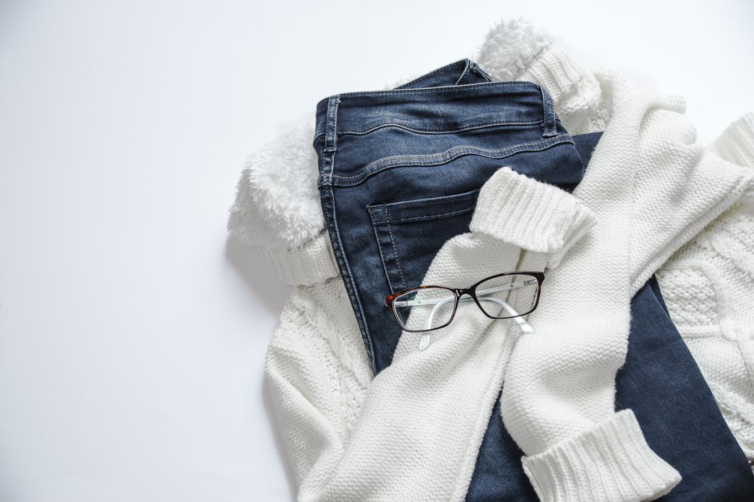 A simple flat lay of black eyeglasses, a white jacket, and blue denim on a plain background