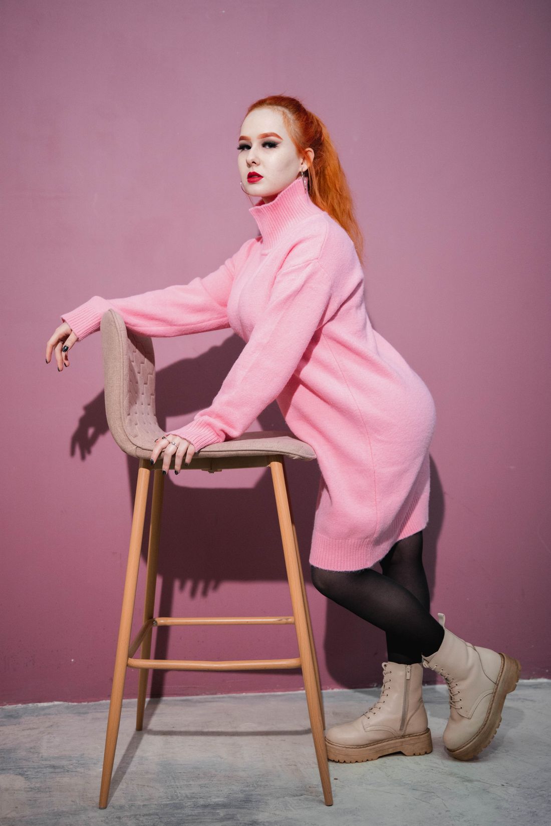 A woman in a solid pink sweater dress leaning against a wooden stool, shot three-quarter length against a plain backdrop.