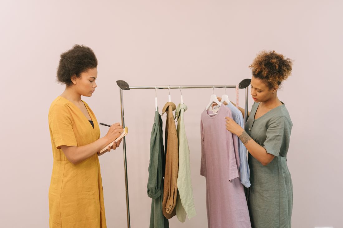 Two people beside a clothing rack holding muted, tonally coordinated garments in sage, tan, and dusty mauve, one taking notes.