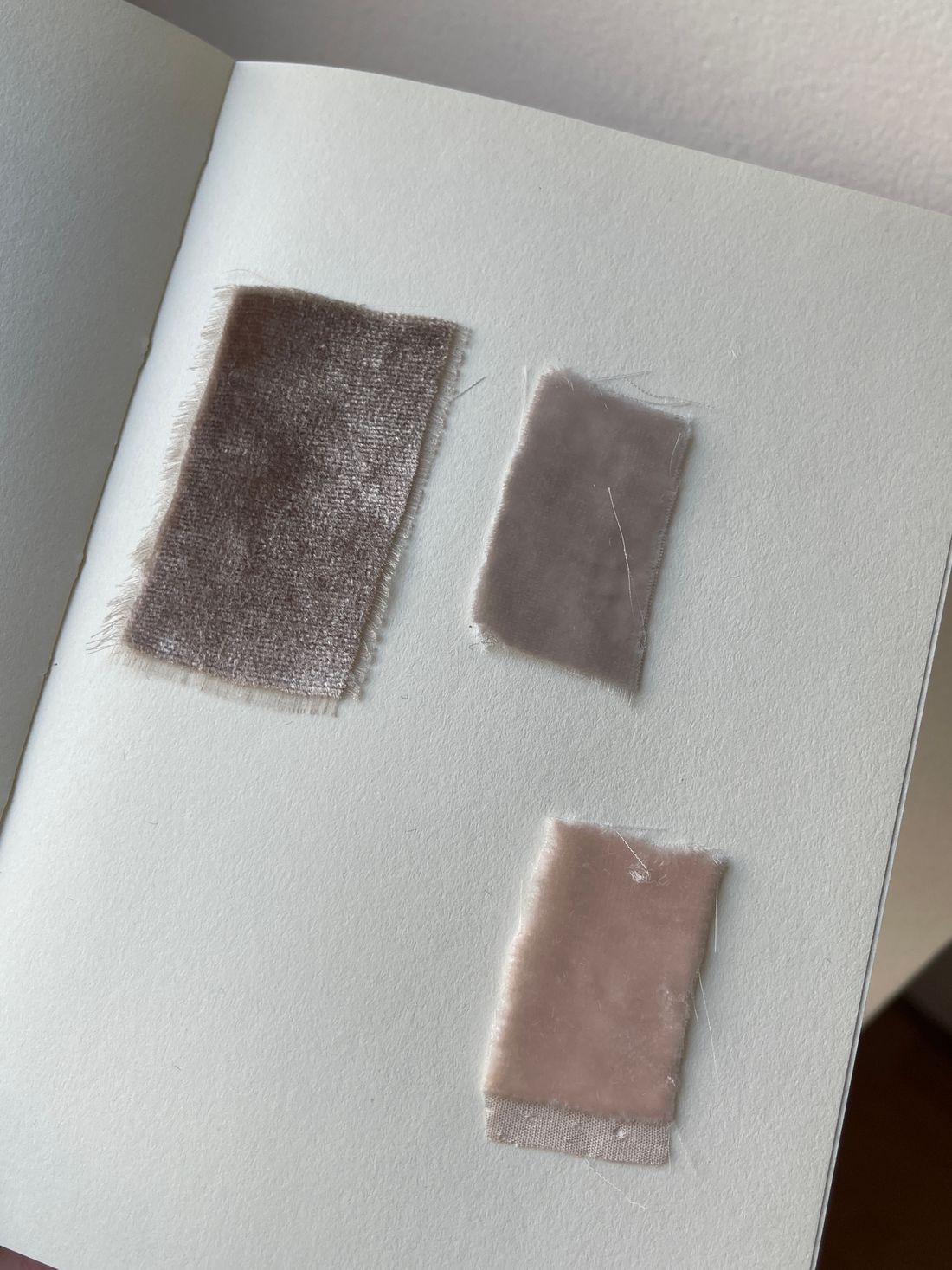 A top-down view of three fabric swatches with raw edges laid on an open notebook page, ranging from taupe to soft pink.