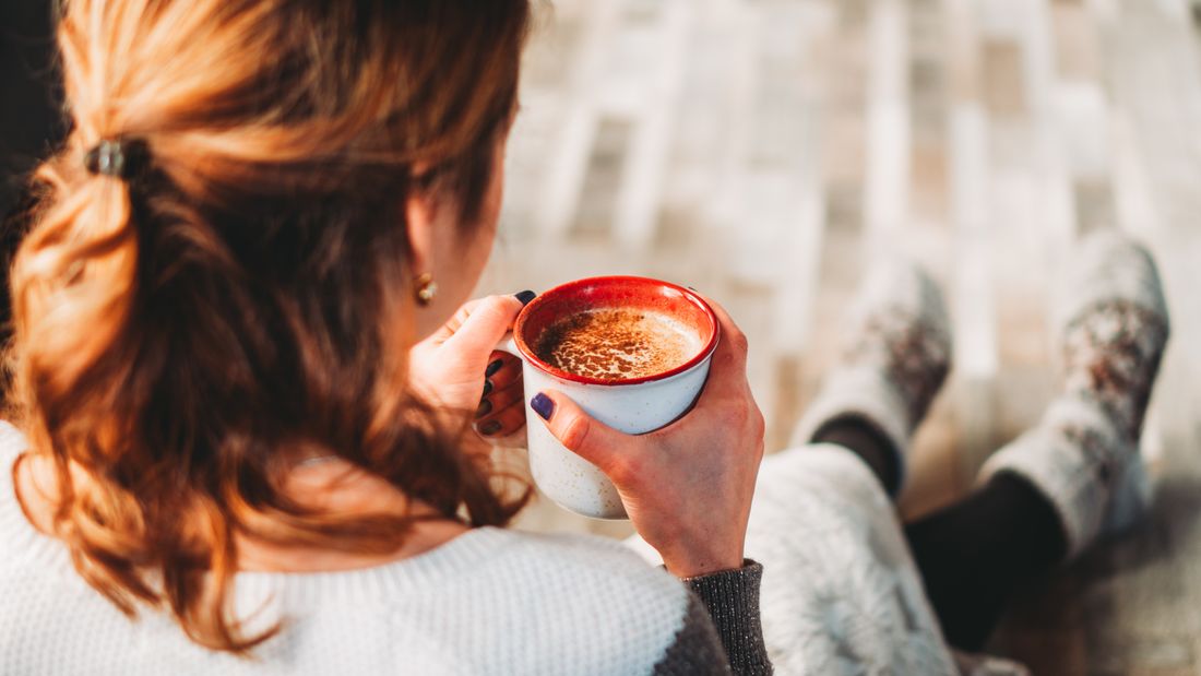 A seated person in a plain cream sweater seen from behind, holding a plain white and red mug indoors