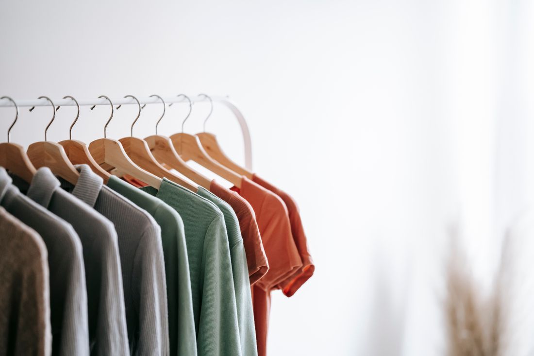 A garment rail in a bright room holding a small number of sage green, terracotta, and gray tops on wooden hangers, seen at a slight angle