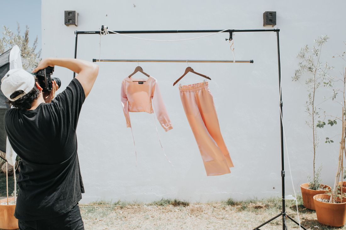 A stylist photographing a peach top and pants on a portable clothing rack set up against an outdoor wall
