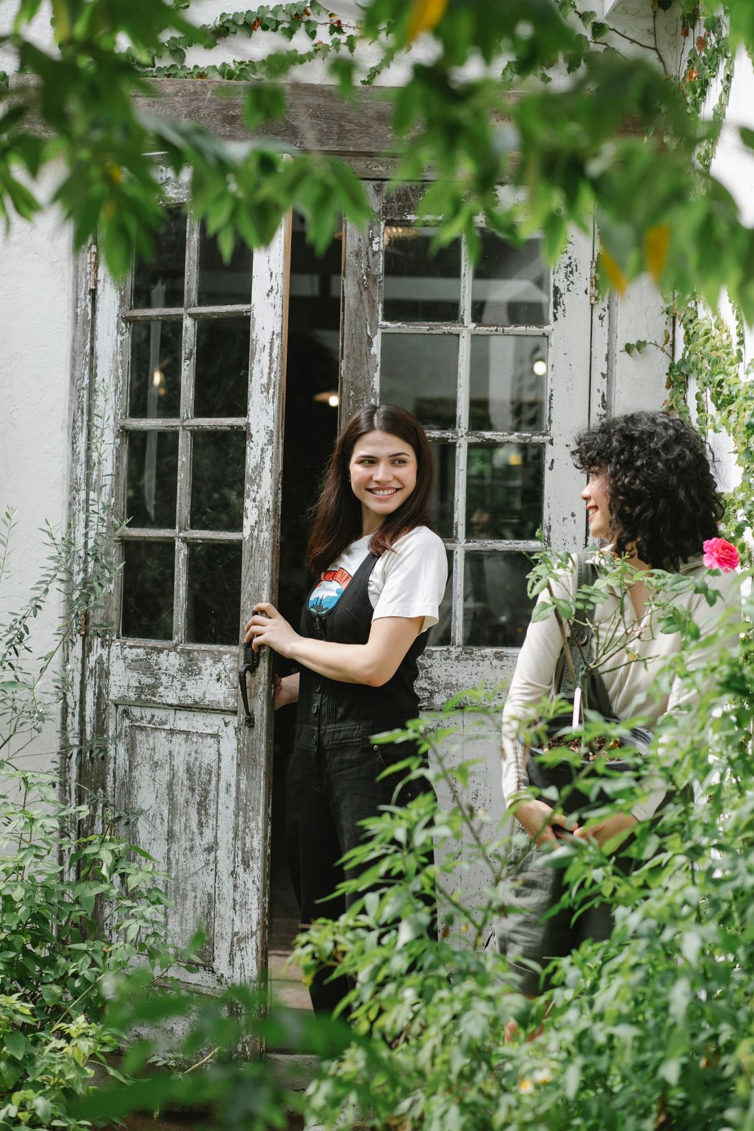 Two women standing at an open weathered wooden door surrounded by garden greenery