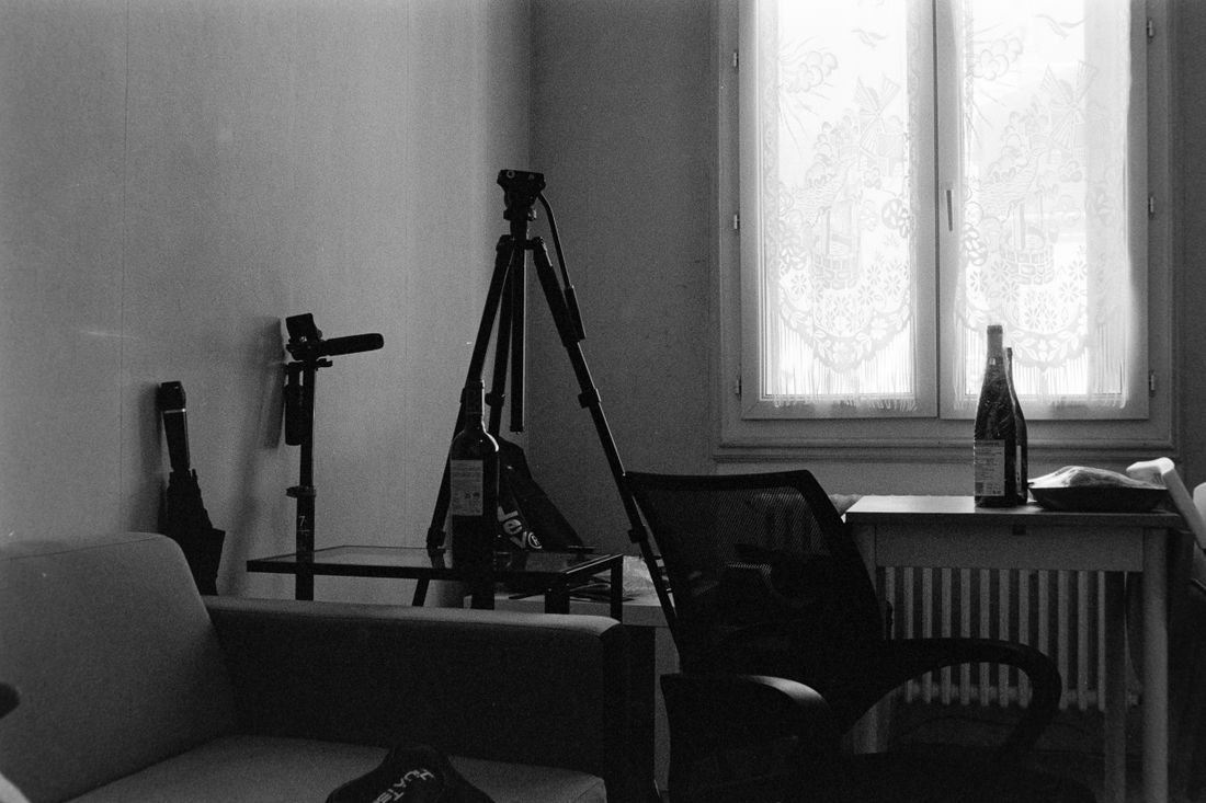 A black-and-white view of a small studio corner with a camera tripod, a chair, and window light